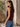 A woman wearing a sleeveless deep plum purple top and blue jeans, standing in profile against a stucco wall and wooden door.