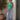 Woman wearing a green tank top and blue jeans standing against a white wall with a plant in the background.