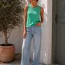 Woman wearing a green tank top and blue jeans standing against a white wall with a plant in the background.