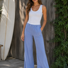 Woman wearing a white tank top and blue pants standing next to a wooden wall and surfboard.