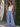 Woman wearing a white tank top and blue pants standing next to a wooden wall and surfboard.