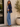 Woman wearing a navy blazer and blue jeans standing against a white wall with a plant in the background.