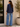 Woman wearing a navy blouse and blue jeans standing against a white wall with a plant in the background.