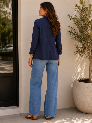 Woman wearing a navy blouse and blue jeans standing against a white wall with a plant in the background.