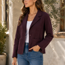 A woman wearing a dark burgundy blazer over a white tank top and blue jeans, standing outdoors against a light-colored stucco wall with green foliage.