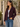 A woman wearing a dark burgundy blazer over a white tank top and blue jeans, standing outdoors against a light-colored stucco wall with green foliage.