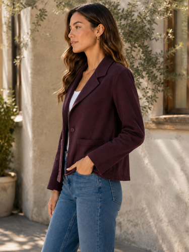 A woman wearing a dark plum purple blazer over a white top and blue jeans, standing outdoors against a textured stucco wall with green foliage.