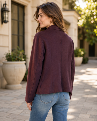 A woman wearing a dark burgundy long-sleeve cropped jacket and blue jeans, photographed from behind in an outdoor courtyard setting.