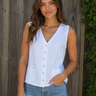 Woman wearing a white sleeveless top with buttons and blue jeans against a wooden background.