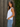 Woman wearing a white sleeveless top and light blue jeans standing against a wooden wall with greenery.