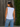 Woman wearing a white sleeveless top and blue jeans standing against a wooden fence with greenery.