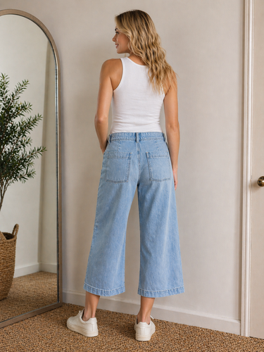 Woman wearing a white tank top and light blue jeans standing in front of a mirror.
