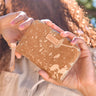 A person holds a zip-around wallet with a tan and beige splatter pattern. The wallet has a small tan leather brand patch that reads Consuela and a light beige zipper pull tab.