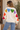 A woman wearing a cardigan with multi-colored hearts across the shoulders down to the arms and blue jeans.