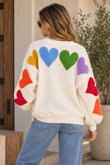 A woman wearing a cardigan with multi-colored hearts across the shoulders down to the arms and blue jeans.