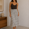 Woman wearing a black top and patterned skirt standing in a room with a wooden dresser and framed picture.