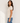A woman wearing a short-sleeve crewneck knit top in a warm off-white/cream color, paired with blue jeans, standing against a plain off-white wall with soft window shadows.