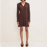 A woman modeling a ribbed knit long-sleeve mini dress in a dark brownish-purple color, paired with matching pointed-toe heels, against a plain off-white studio background.