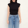 A woman wearing a black sleeveless mock-neck top and light blue jeans, standing against a plain off-white background.