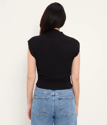 A woman viewed from the back wearing a fitted black sleeveless mock-neck top and light blue jeans against a plain off-white background.
