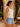 A woman wearing a white textured sleeveless tank top and light blue denim shorts, standing in a sunlit outdoor patio setting with a beige stucco wall, potted plants, and a wicker chair.