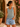 Woman wearing a light blue tank top and denim shorts standing in a sunlit outdoor setting with plants.