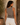 Woman wearing a brown tank top and white pants standing against a beige wall with a plant in the background.