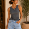 Woman wearing a gray tank top and denim shorts standing against a beige wall with a plant in the background.