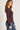 A woman wearing a fitted, long-sleeve crewneck top in a deep plum-brown color, paired with blue jeans, shown in a side-profile view against a plain light background.