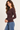 A woman wearing a fitted, long-sleeve crewneck top in a deep plum-brown color, paired with blue jeans, shown in a side-profile view against a plain light background.