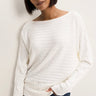 A woman wearing a white, long-sleeve knit sweater with a wide boat neckline and horizontal ribbed texture, shown against a plain off-white background.