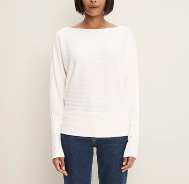 A woman wearing a white long-sleeve knit top with a wide boat neckline and textured vertical ribbing, paired with dark blue jeans, against a plain light beige background.