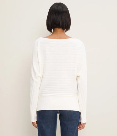 A woman viewed from the back wearing a cream-colored, long-sleeved ribbed knit top and dark blue jeans, set against a plain off-white background.