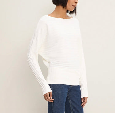 A woman wearing a white long-sleeve knit sweater with textured vertical pleats and ribbed cuffs, paired with blue jeans, against a plain off-white studio background.