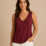 A woman modeling a sleeveless V-neck tank top in burgundy, paired with white pants, against a solid beige background.