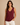 A woman modeling a sleeveless V-neck tank top in burgundy, paired with white pants, against a solid beige background.