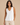 A woman modeling a white sleeveless V-neck tank top with light beige pants, standing against a solid beige studio background.