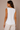 A woman shown from the back wearing a white sleeveless raglan top and light beige pants, standing against a solid warm beige studio background.
