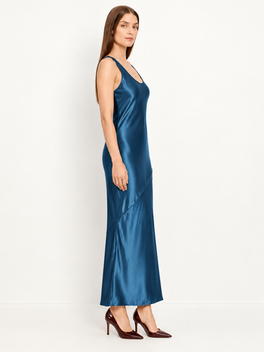 A woman modeling a satin slip maxi dress in a deep teal blue color, paired with dark brown pointed-toe heels, against a plain white studio background.