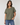 A woman wearing a relaxed-fit, short-sleeve crewneck t-shirt in olive green, paired with blue jeans, standing against a light gray studio background.