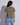 A woman viewed from behind wearing an olive green short-sleeve t-shirt and light blue jeans, standing against a light gray studio background.