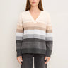 A woman wearing a long-sleeve V-neck knit sweater with horizontal stripes in cream, beige, taupe, light gray, medium gray, and charcoal, paired with dark gray jeans against a plain off-white background.