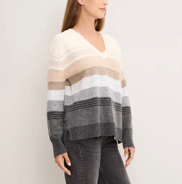 A woman wearing a knit V-neck sweater with horizontal stripes in cream, beige, white, light gray, medium gray, and dark charcoal gray, paired with dark gray jeans against a plain light gray background.