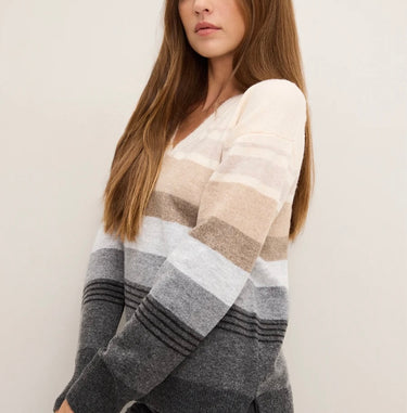 A woman wearing a fuzzy knit sweater with horizontal stripes in cream, beige, taupe, light gray, medium gray, and dark charcoal gray, shown from the chest up against a plain off-white background.