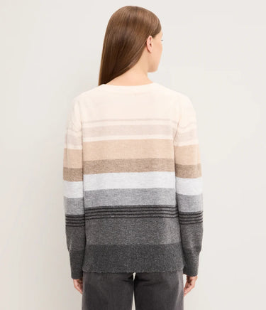 A woman viewed from the back wearing a striped knit sweater with horizontal bands in cream, beige, light gray, medium gray, and dark charcoal gray, paired with dark gray pants, against a plain light gray studio background.