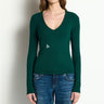 A woman wearing a dark green long-sleeve V-neck knit top with a small white geometric logo on the chest, paired with blue jeans, standing against a plain off-white wall.