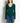 A woman wearing a dark green long-sleeve V-neck knit top with a small white geometric logo on the chest, paired with blue jeans, standing against a plain off-white wall.