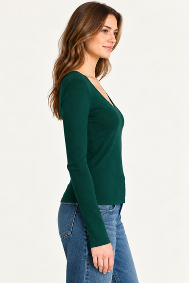 A woman wearing a dark green long-sleeve V-neck top, paired with blue jeans, standing against a plain off-white wall.