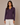 A woman modeling a long-sleeve button-up shirt in a deep purple color, paired with white pants, against a neutral beige studio background.