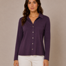 A woman modeling a long-sleeve button-up shirt in a deep purple color, paired with white pants, against a neutral beige studio background.